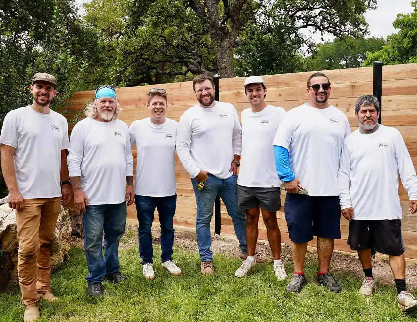 A group of eight men wearing matching white long-sleeve shirts standing outdoors, smiling at the camera with a wooden fence in the background.