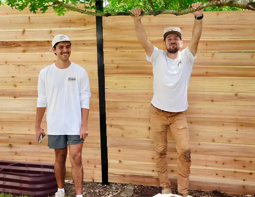 Two men standing next to a wooden fence, one hanging from a branch while the other smiles beside him. Both are wearing casual clothing and hats.